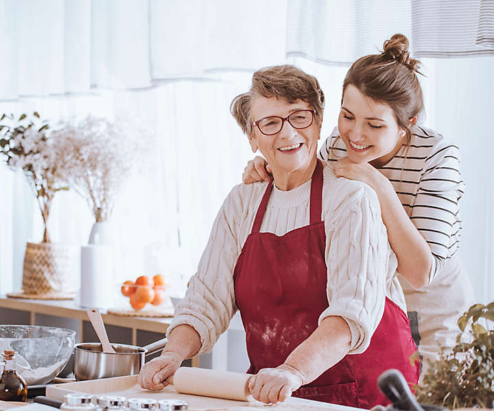 Young woman looking over the shoulder of her grandmother, who is rolling out pie dough