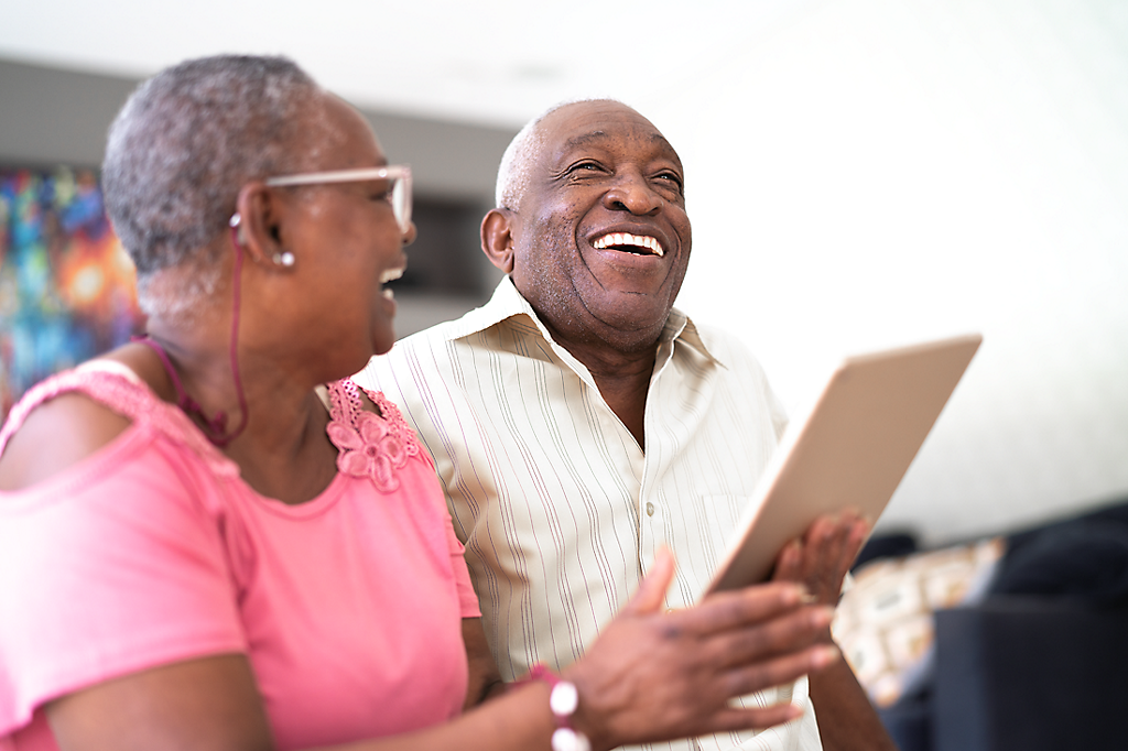 A mature couple laughing and smiling at something on a tablet.