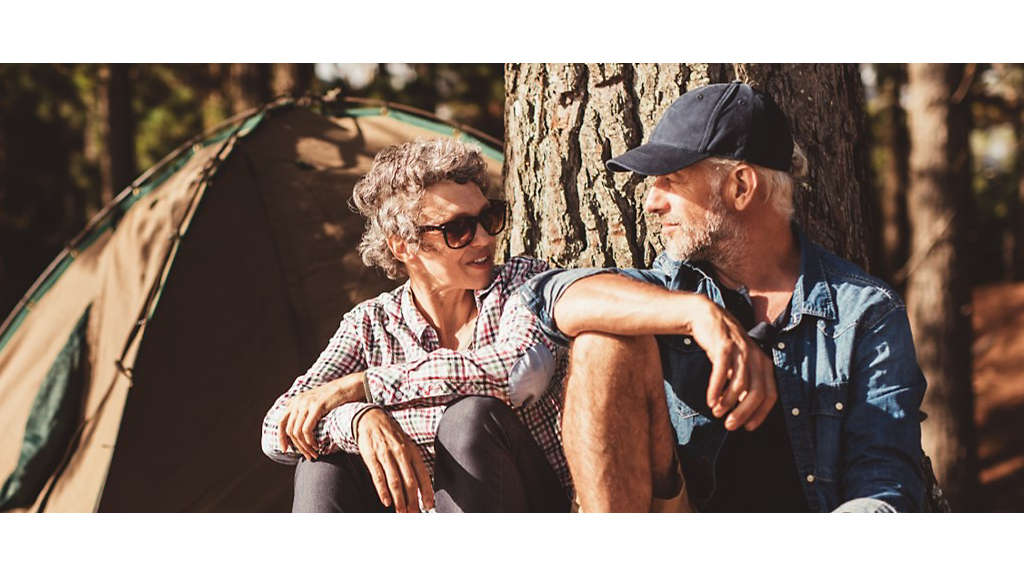 A mature couple sitting under a tree outside a camping tent.