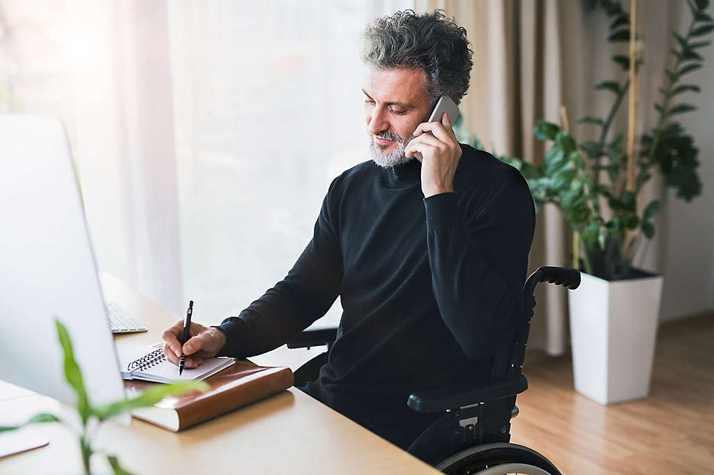 A man in a wheelchair sitting at a desk and talking on a cell phone.